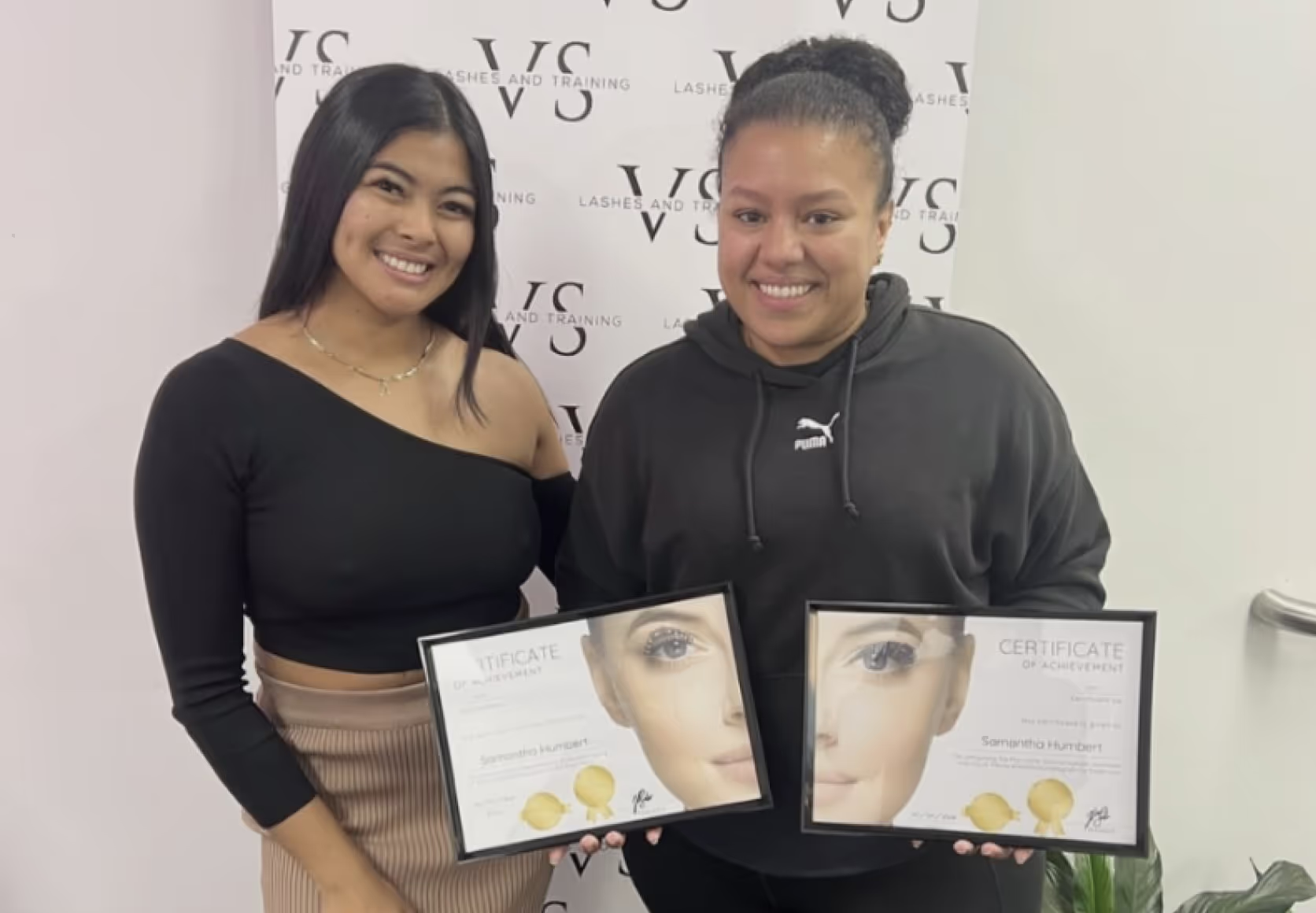 Trainer and student holding framed lash certification diplomas in beauty studio setting.
