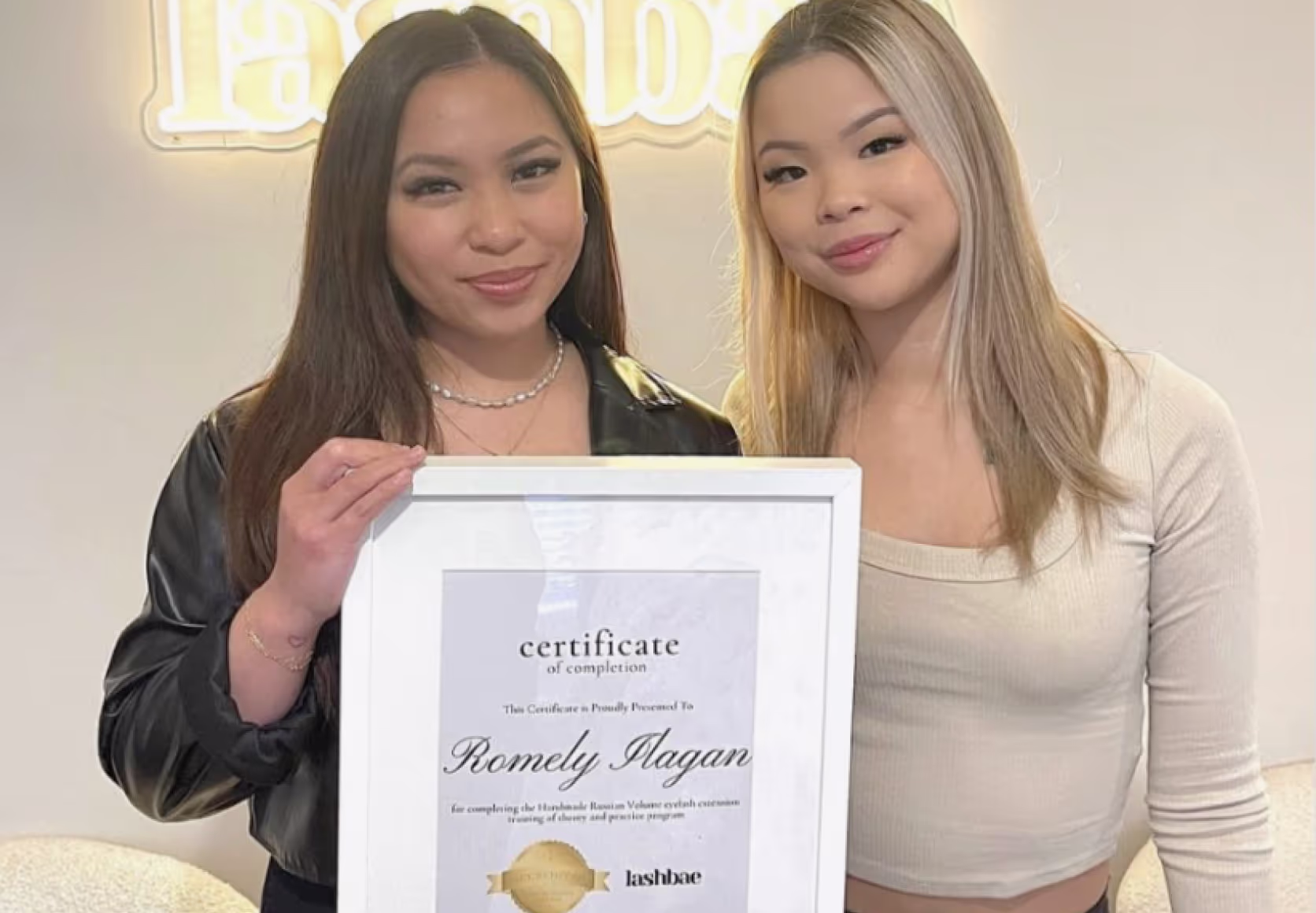 Trainer and student holding framed lash certification diploma in beauty studio setting.