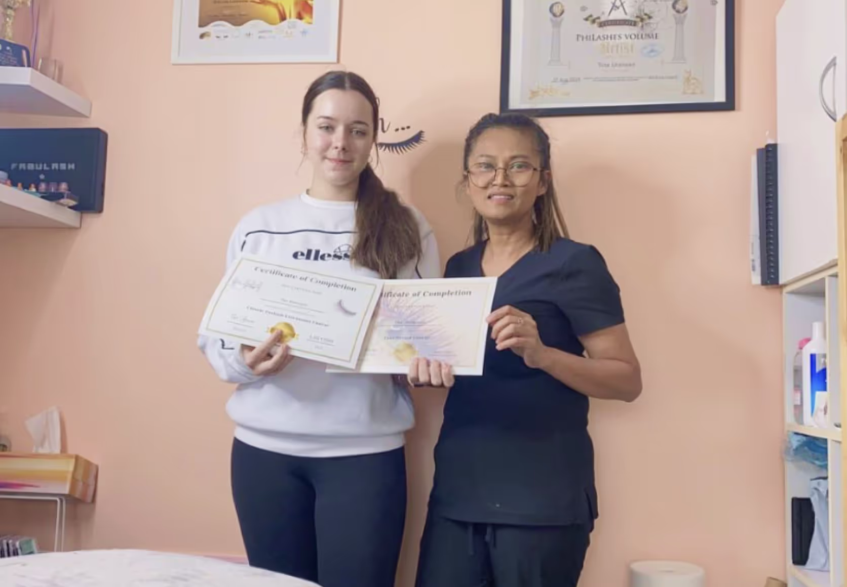 Trainer and student holding lash certification diplomas in beauty studio setting.