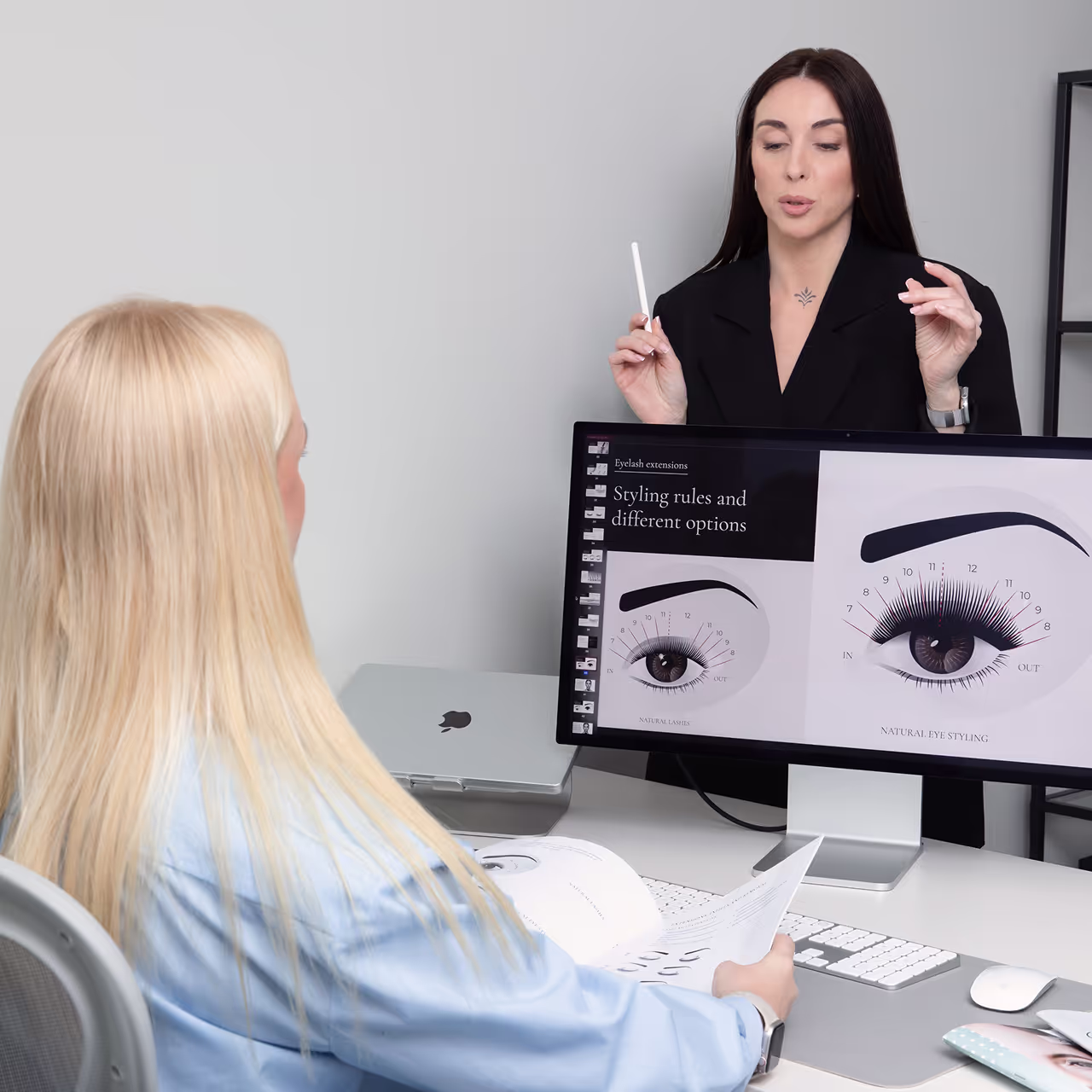 Lash educator teaching a student with eyelash styling diagrams on screen during a certification course.