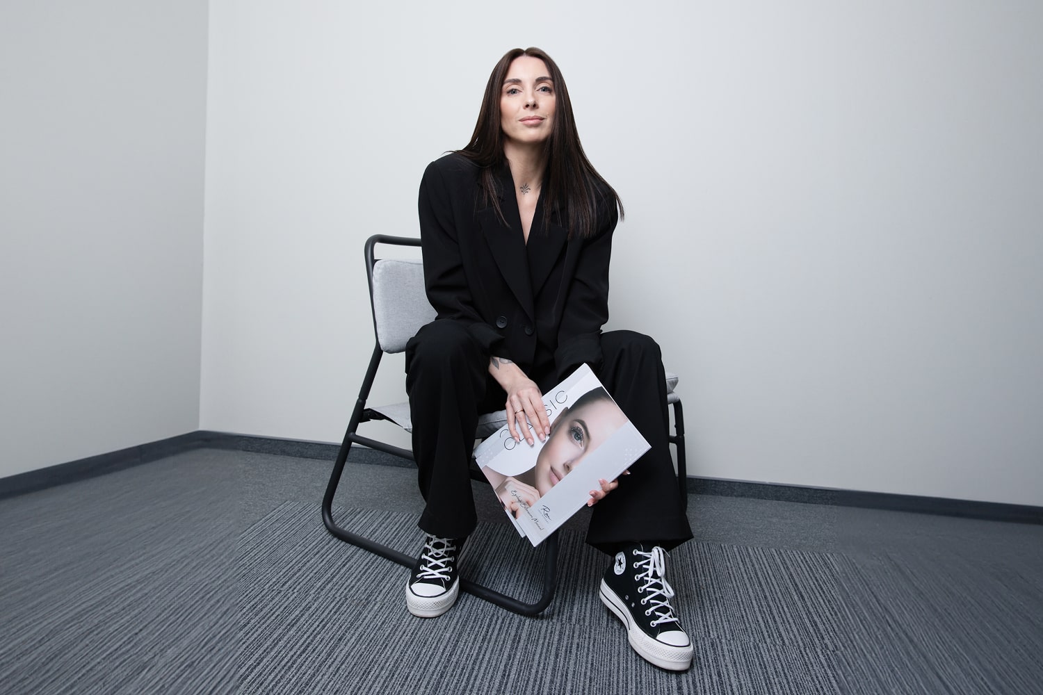 A woman sitting on a chair, holding eyelash extension course materials in their hands, wearing a black suit and Converse sneakers, with a minimalist background.