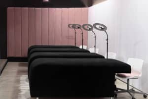 An image of a modern lash salon interior with a row of black, flat reclining salon beds aligned neatly. Behind the beds is a pink velvet paneled wall. Each bed is accompanied by a white chair and a black adjustable salon lamp with a circular light and a magnifying glass in the center. The salon floor is wooden, and the overall aesthetic is clean and professional, with a touch of soft color from the pink wall.
